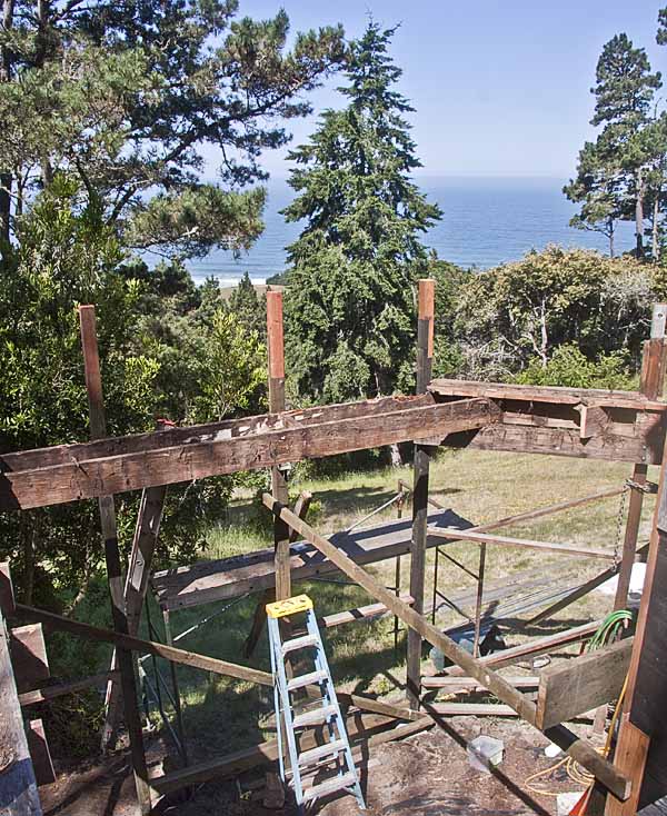 The framework of the old deck with a ladder and a view of the ocean beyond some trees.